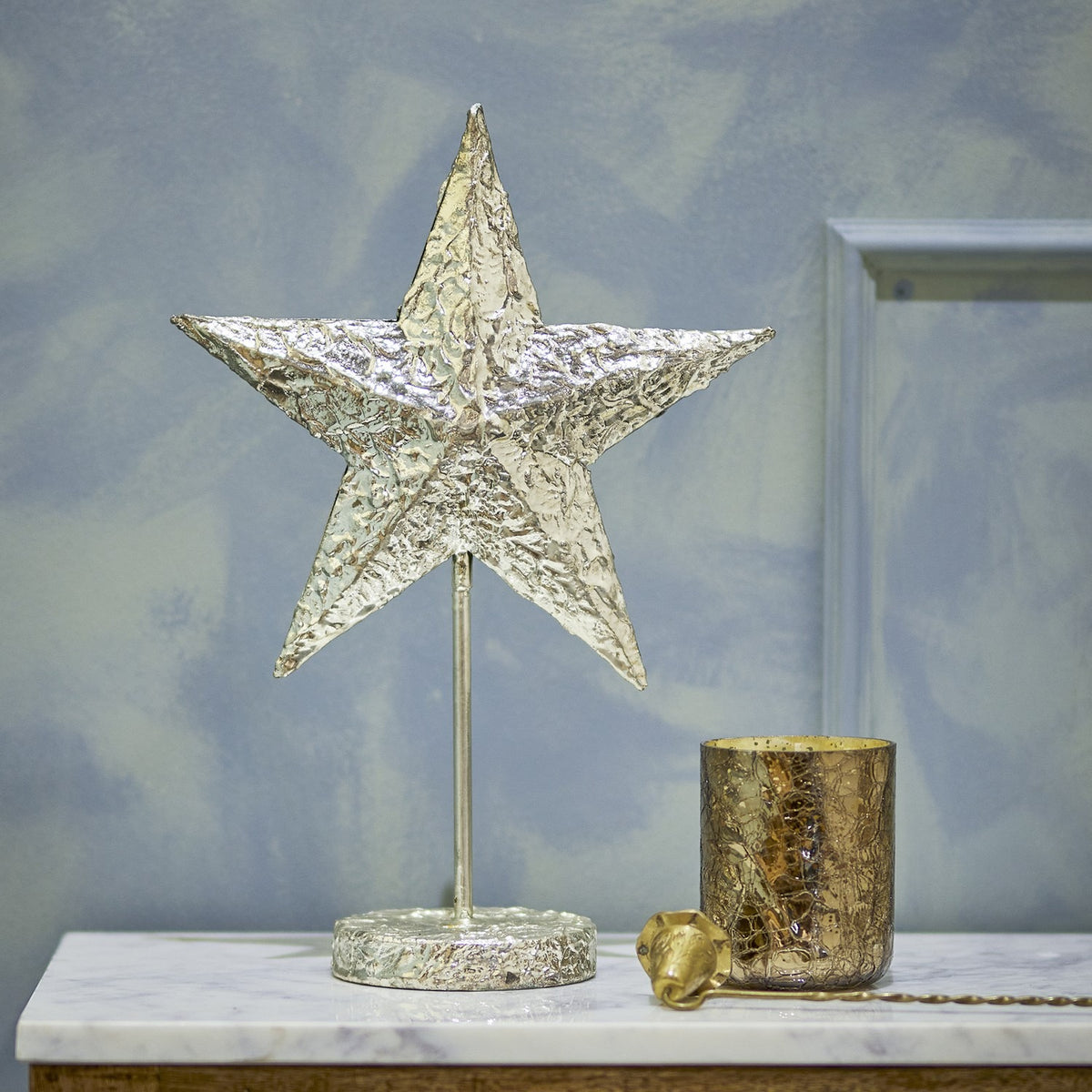 Nilos Star Iron Christmas Decoration in brass, a textured five-pointed star on a slim stem and round base, shown on a marble shelf beside a mercury glass votive.
