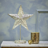 Nilos Star Iron Christmas Decoration in brass, a textured five-pointed star on a slim stem and round base, shown on a marble shelf beside a mercury glass votive.