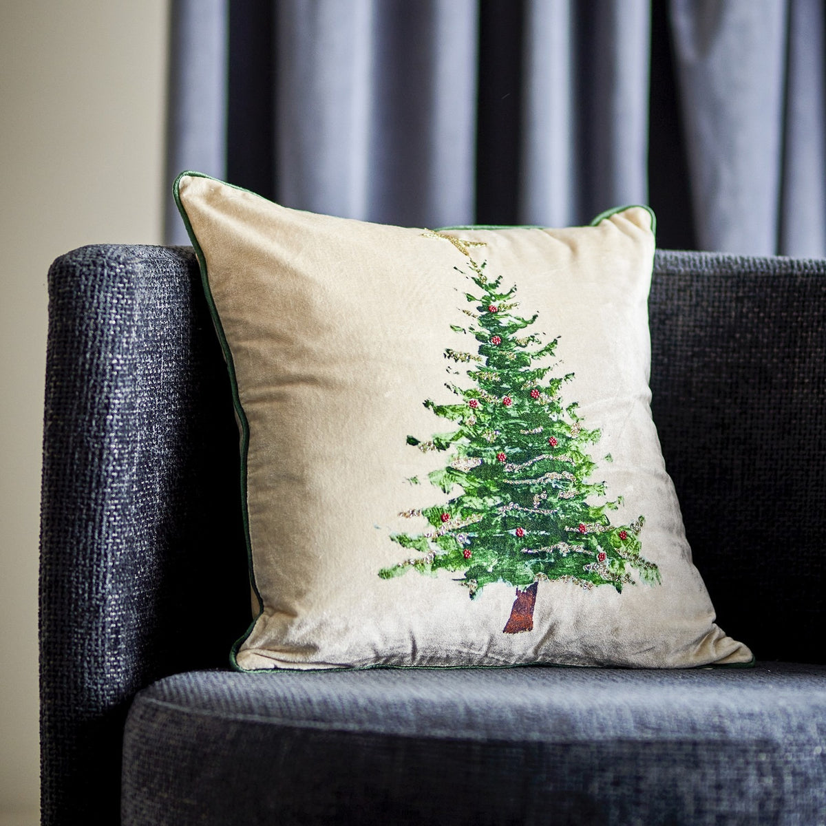 Evelin Cotton Christmas Cushion in cream with a painted green Christmas tree design, green piped edge, shown on a dark grey sofa against striped curtains.