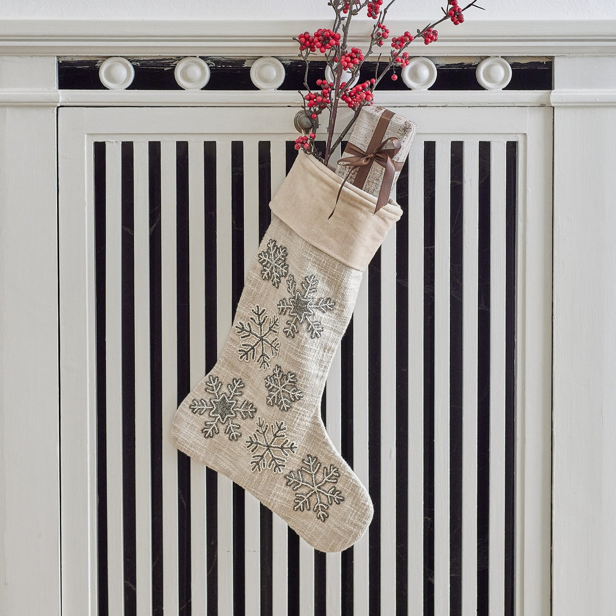 Ahsan Snowflake Cotton Christmas stocking in beige woven fabric with a folded cuff, beaded snowflake embroidery down the front, hanging filled with a wrapped gift and red berry sprigs.