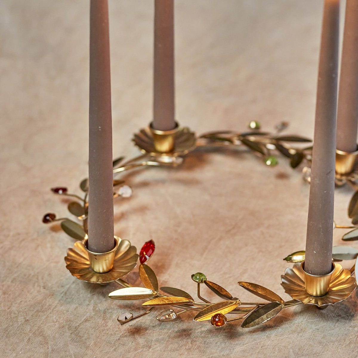 Gold iron Advent candle holder wreath with four flower-shaped cups holding taupe candles, accented by leaf sprigs and coloured faceted berry details.