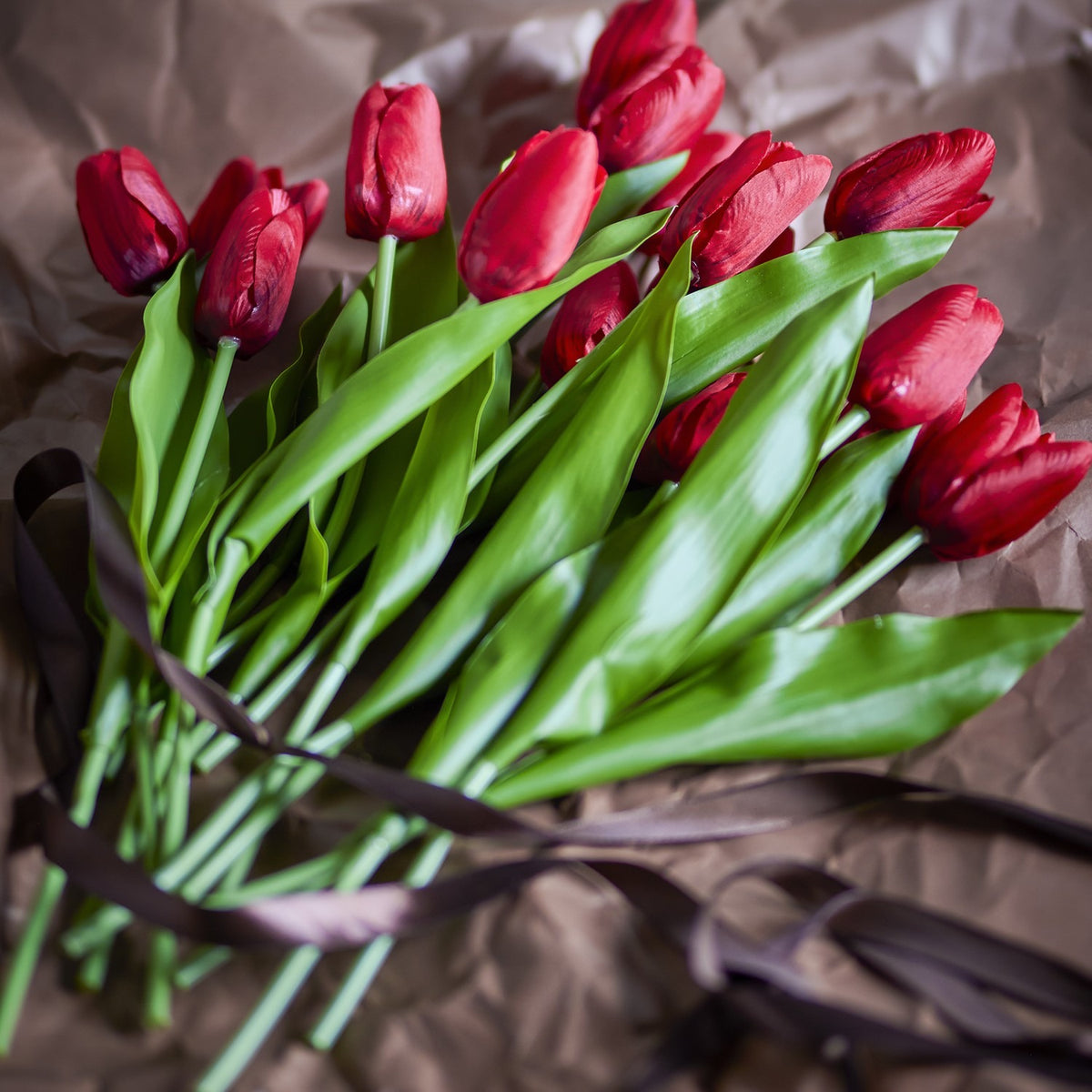 Artificial tulip bouquet in red with cupped flower heads, long green stems and broad leaves, tied with brown ribbon on crumpled paper backdrop.