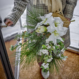 Kristin Artificial Christmas Bouquet in white, held in the crook of an arm by a doorway, showing large trumpet blooms, pine fronds, small cones and white berry clusters bound with twine.