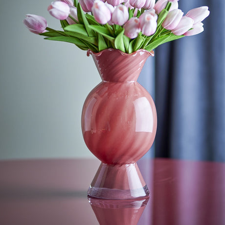 Rajan Wavy Glass Vase 29cm in pink, with a frilled wavy rim, round swirled belly and conical foot, holding pink tulips on a reflective pink surface.