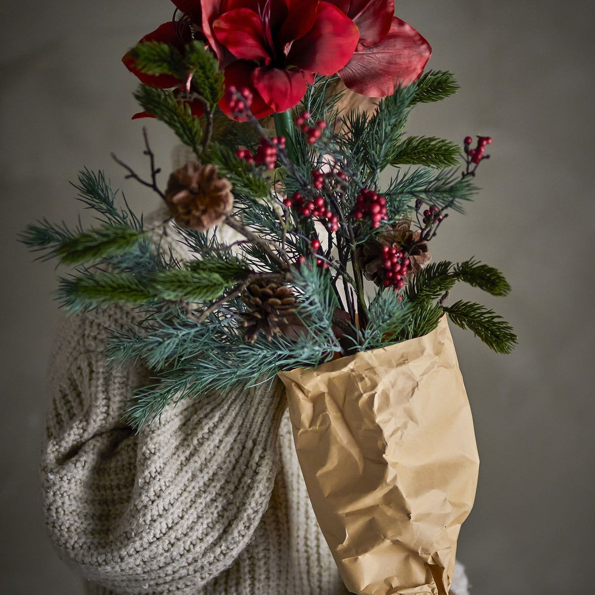 Rubia Artificial Christmas Bouquet in red, with large faux flowers, pine sprigs, pinecones and berries, held in kraft paper wrap against a knitted sweater.