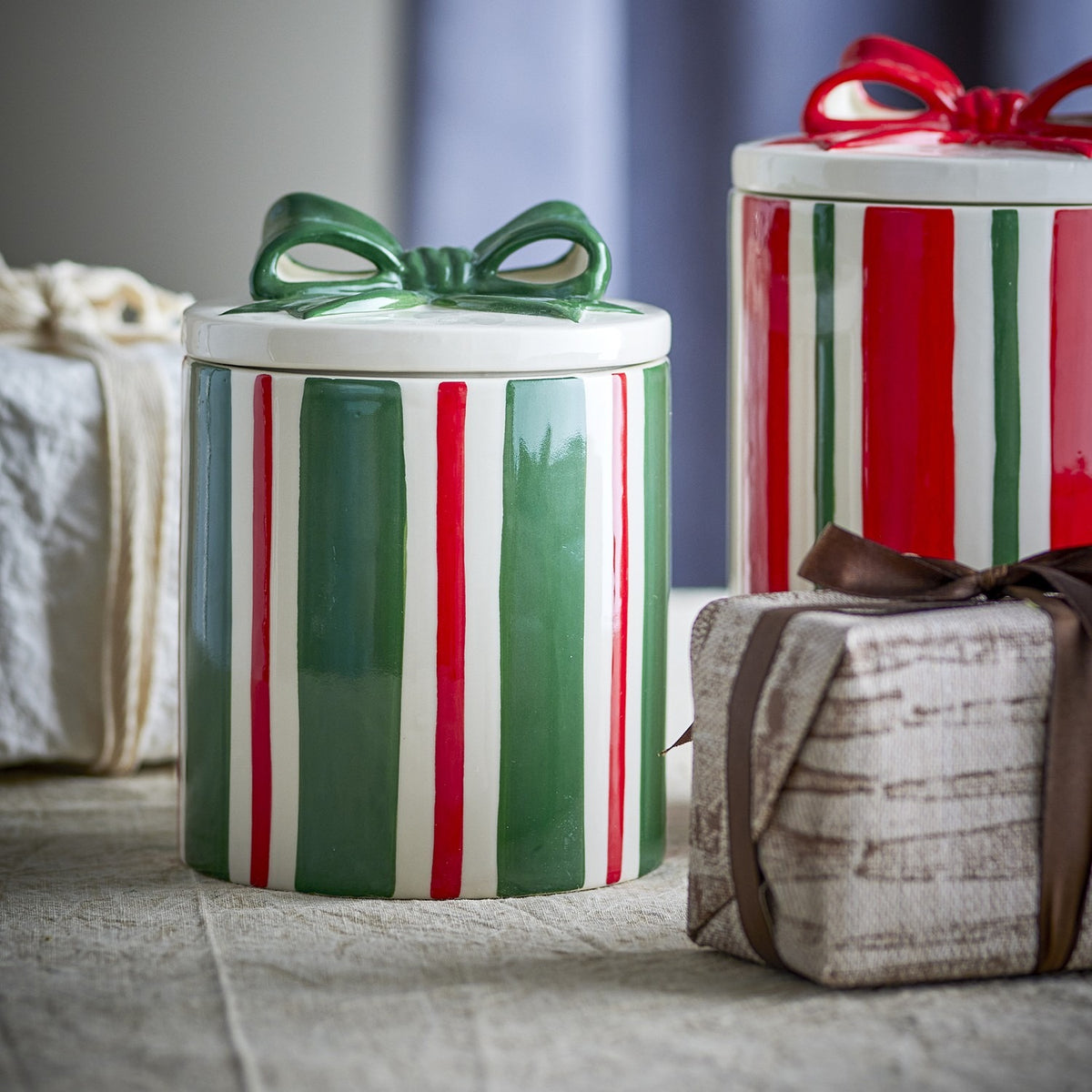 Marnia ceramic Christmas storage jar with green and red striped body, white lid, and green bow, shown on a table beside gift boxes.