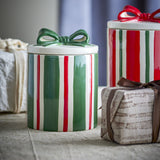 Marnia ceramic Christmas storage jar with green and red striped body, white lid, and green bow, shown on a table beside gift boxes.