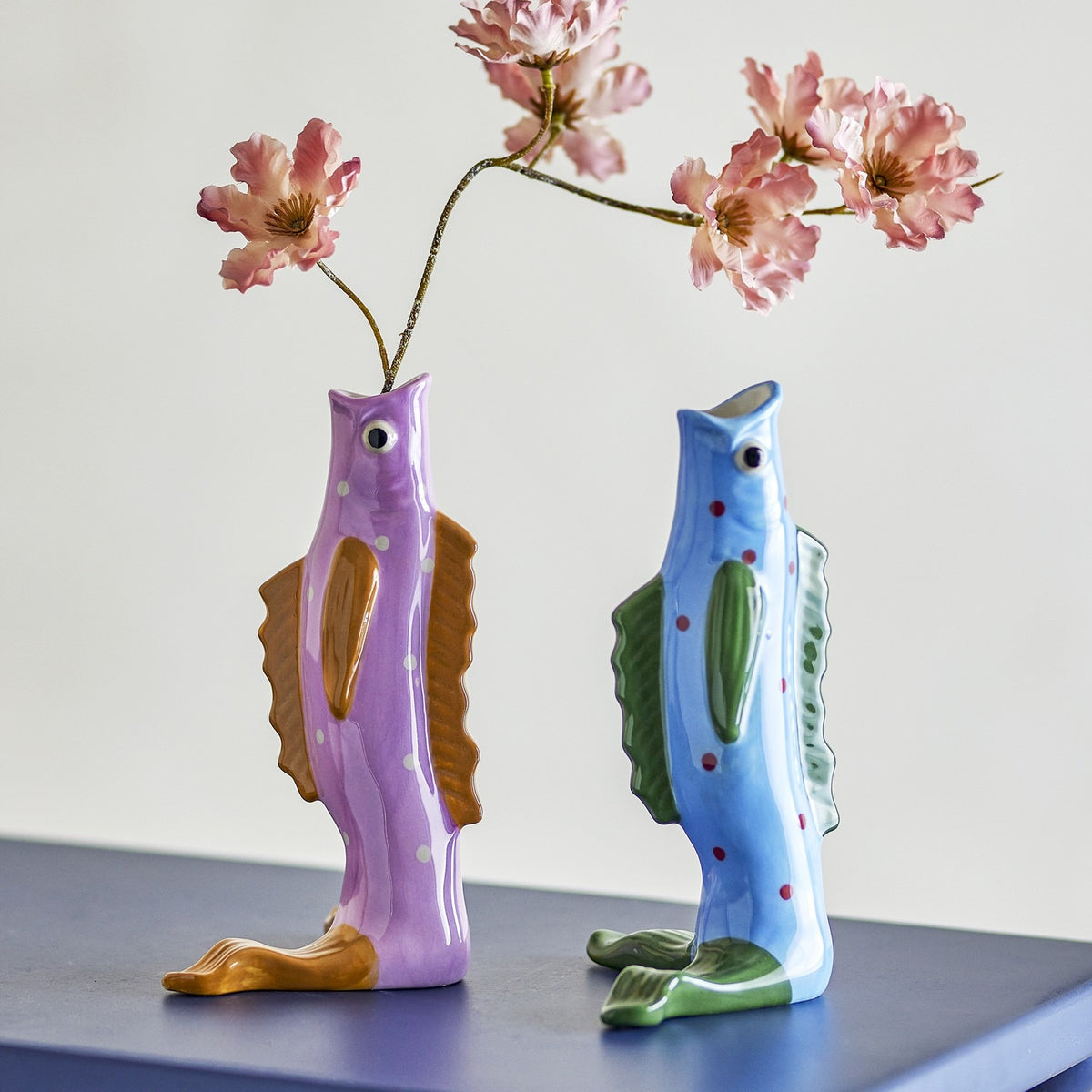 Mako Fish Stoneware Vases 19cm Set of 2 in purple and blue, upright fish-shaped with painted eyes and ridged fins, holding pink blossom branches on a dark surface.