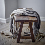 Hamisha Grey Throw draped over a dark wood stool, showing fuzzy multicoloured block-check fabric and long chunky fringe, in a rustic room setting.
