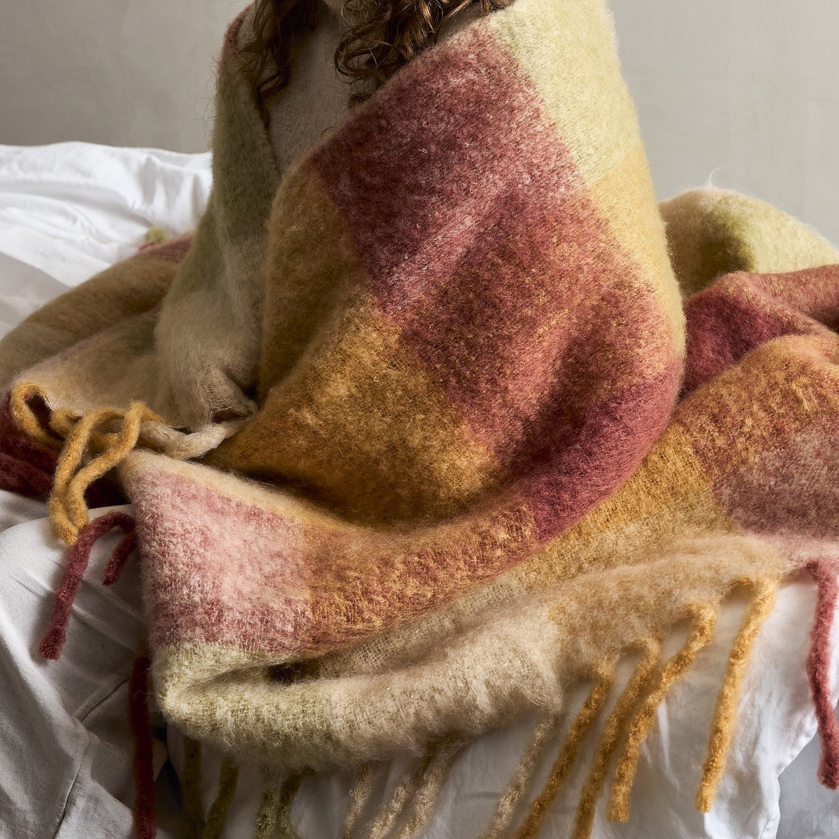 Hamisha Throw - Multi, a large checked woven throw in pink, gold, and green tones with a chunky twisted fringe, draped over a bed.