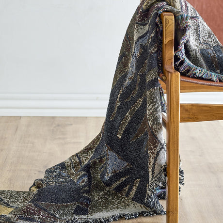 Rahul Cotton Throw in Multi draped over a wooden armchair, showing a woven abstract pattern in dark and neutral tones with a loose multicoloured fringe.