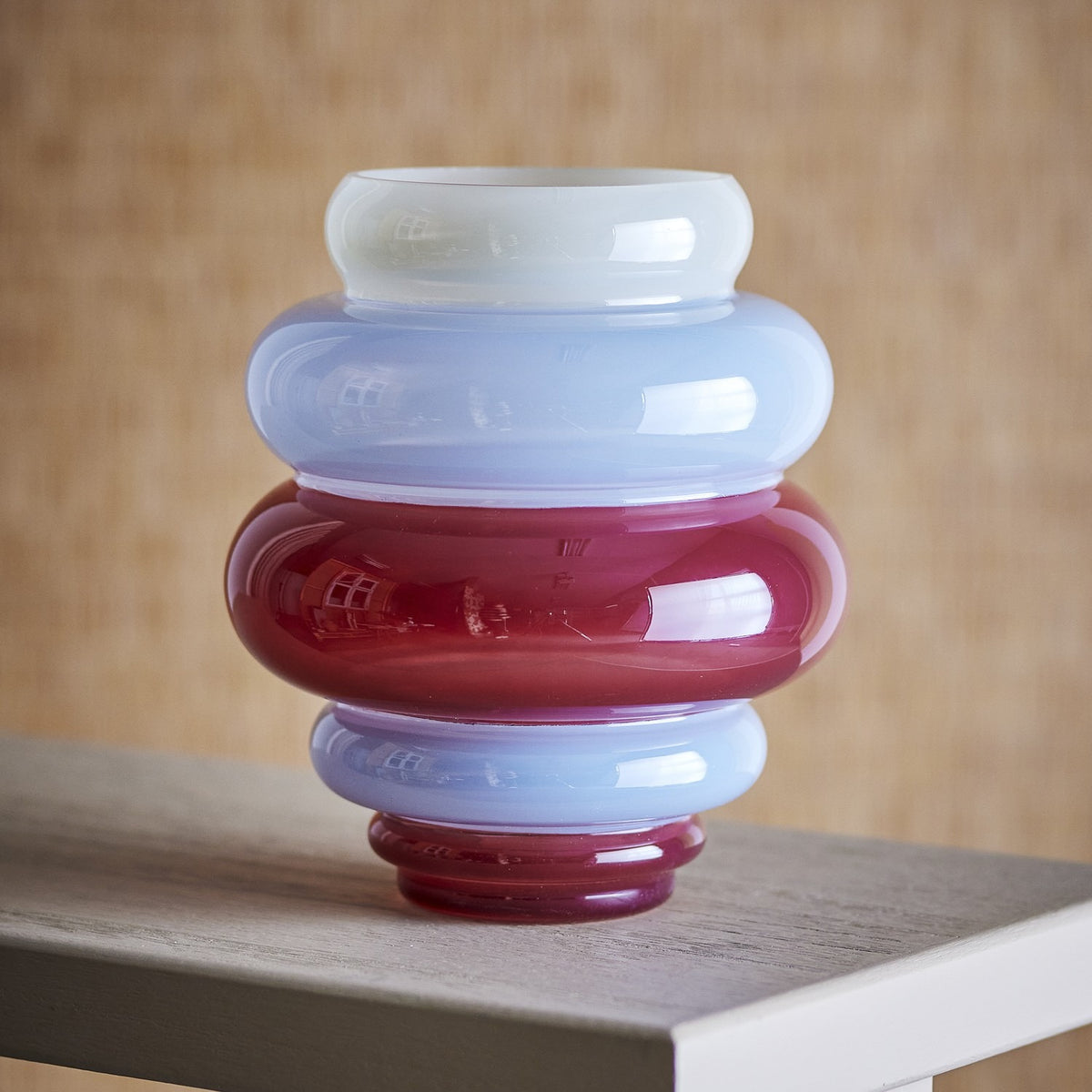 Annhelene Glass Vase 24cm in Multi, featuring stacked rounded tiers in white, blue, and deep red glass with a narrow top ring, shown on a wooden surface against a woven backdrop.