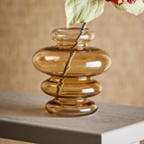 Kayn Glass Vase 19.5cm in Amber, with stacked rounded tiers from neck to base, shown on a tabletop with a leafy stem and warm beige background.