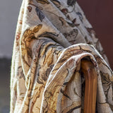 Yoti Floral Cotton Throw in multicoloured botanical print, draped over a wooden armchair frame showing the textured weave and flowing fabric folds.