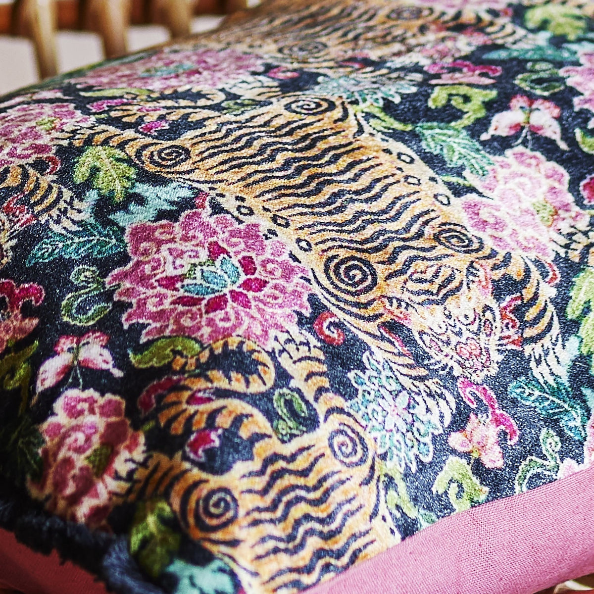 Lester Cotton Cushion in Multi, a rectangular fabric cushion with a dense printed pattern of stylised tigers, blooms and leaves in pink, gold and teal, finished with a pink fabric border.