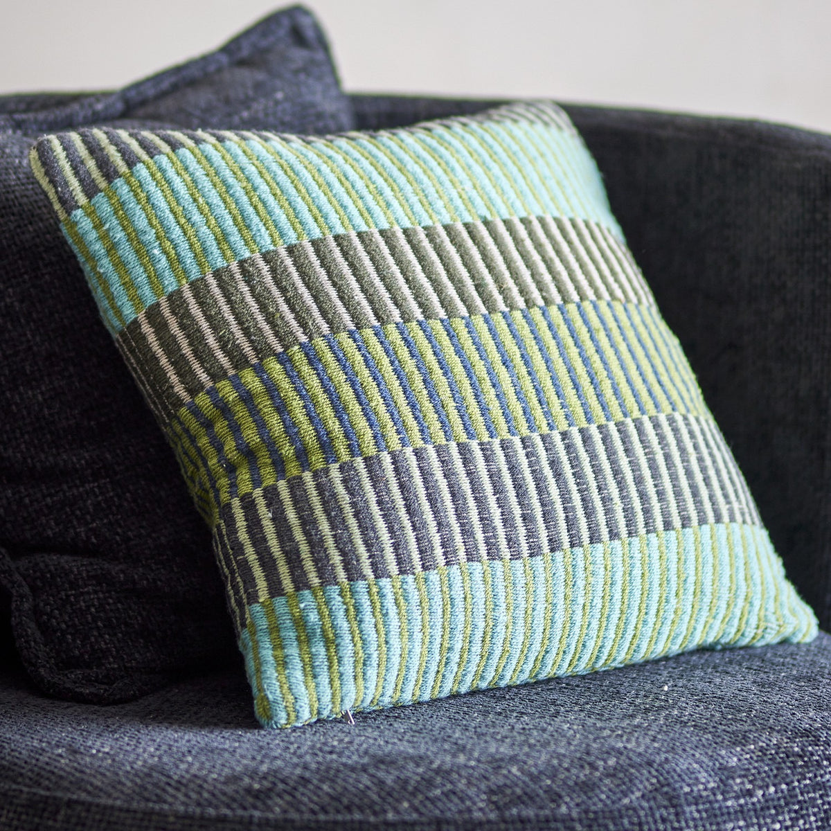 Isnel Green Striped Cushion in multicoloured horizontal stripes of green, blue and grey, woven fabric cover, shown resting on a dark grey sofa.