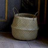 Floria Seagrass Basket in natural, a round belly-shaped storage basket with two-tone zigzag weave and twisted handles, shown against a dark wooden backdrop.