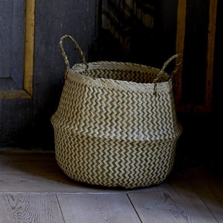 Floria Seagrass Basket in natural, a round belly-shaped storage basket with two-tone zigzag weave and twisted handles, shown against a dark wooden backdrop.