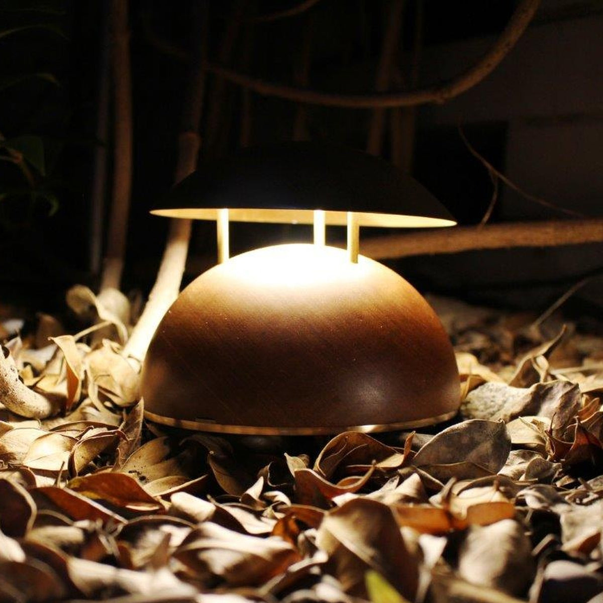 Bell LED rechargeable outdoor table lamp with half-round wooden base and brass-coloured domed metal shade on three rods, glowing warm white among autumn leaves at night.