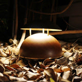 Bell LED rechargeable outdoor table lamp with half-round wooden base and brass-coloured domed metal shade on three rods, glowing warm white among autumn leaves at night.