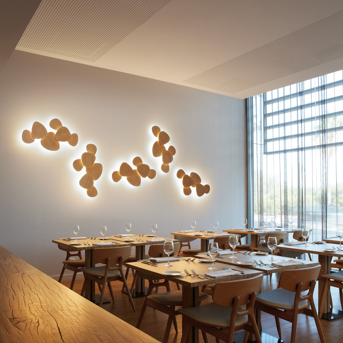 Tria 6 Light Wood LED Wall Light with clustered pebble-shaped wood panels casting warm backlit glow, shown mounted above dining tables in a restaurant setting.