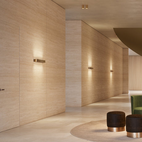 Toy R LED Wall Light, 29.7cm bronze-finish bar fixtures mounted along a travertine corridor, casting warm uplight and downlight on the stone wall.