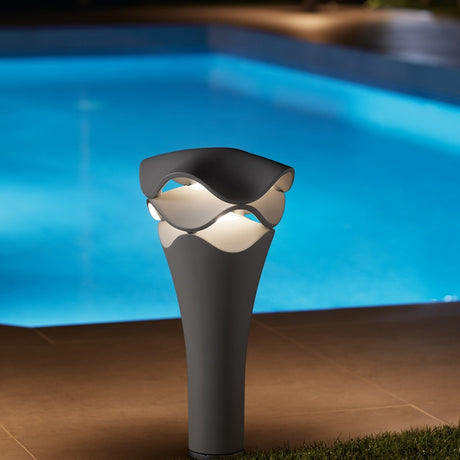 Cornet B/52 cone LED bollard light in stone grey, with wavy layered top emitting warm white glow, shown poolside at night on a patio.