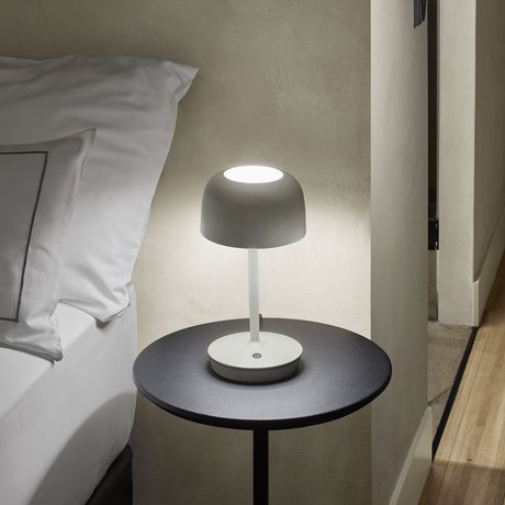 Bol M/37 Dome LED Table Lamp in grey, with a rounded dome shade, slim stem and round base, lit on a bedside table beside white pillows.