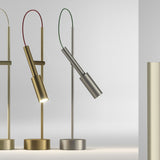 Tubino Aluminium LED Table Lamp in brushed titanium finish, slim stem with angled cylindrical head and looped green cable, round base, shown beside brass versions on a light grey background.