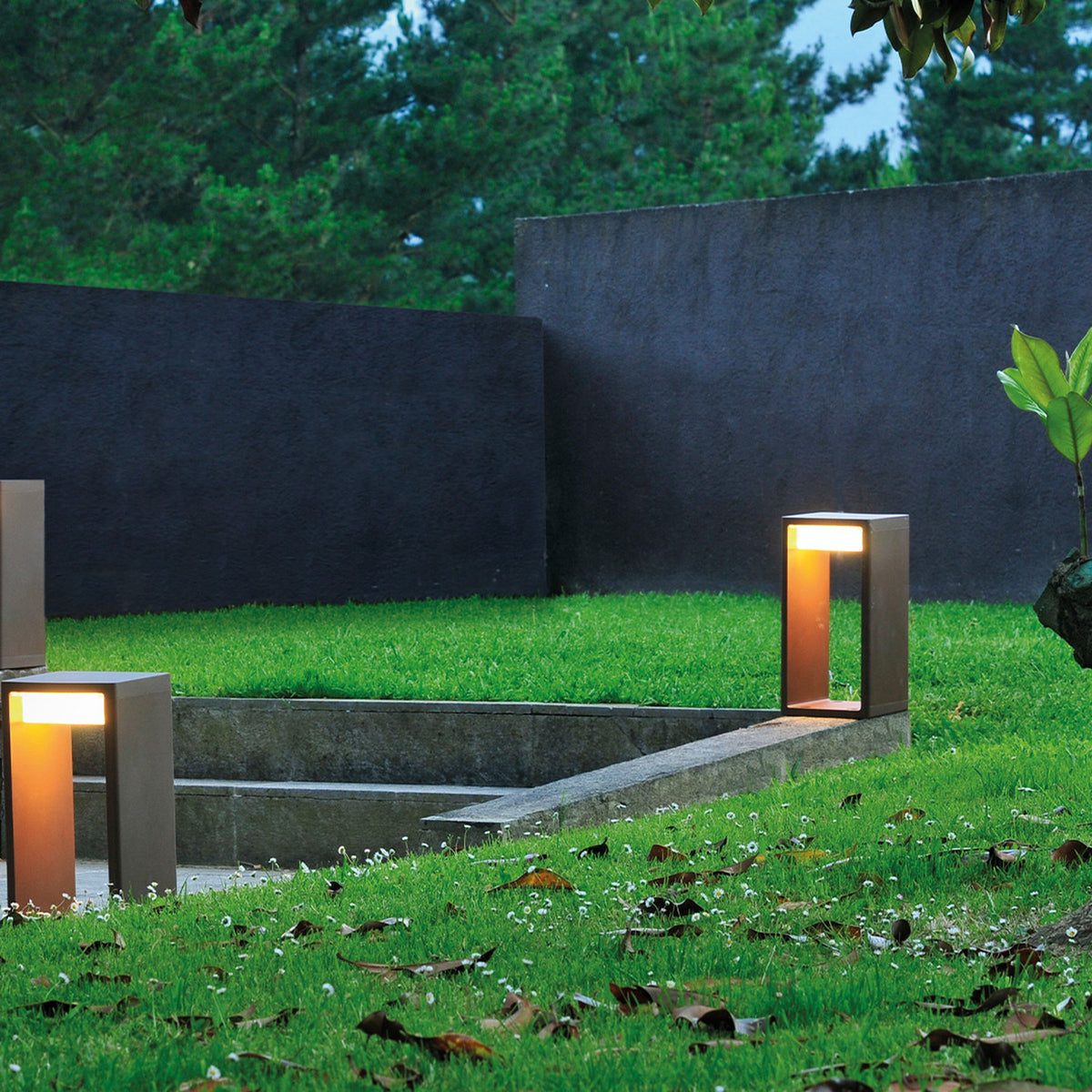Frame M Glass LED Bollard Light in a bronze finish, an open rectangular frame fixture glowing warm white on a lawn beside steps and a dark rendered garden wall.