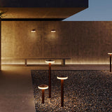 Venexia Glass LED Outdoor Floor Lamp 70cm in Corten, multiple bollard lights with round glass-edged discs glowing warm white on a gravel path beside a concrete courtyard wall at dusk.