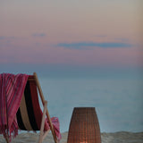 Amphora Woven Linear LED Outdoor Floor Lamp in beige, tapering woven shade glowing warm white beside a striped deckchair and throw on a beach at dusk.