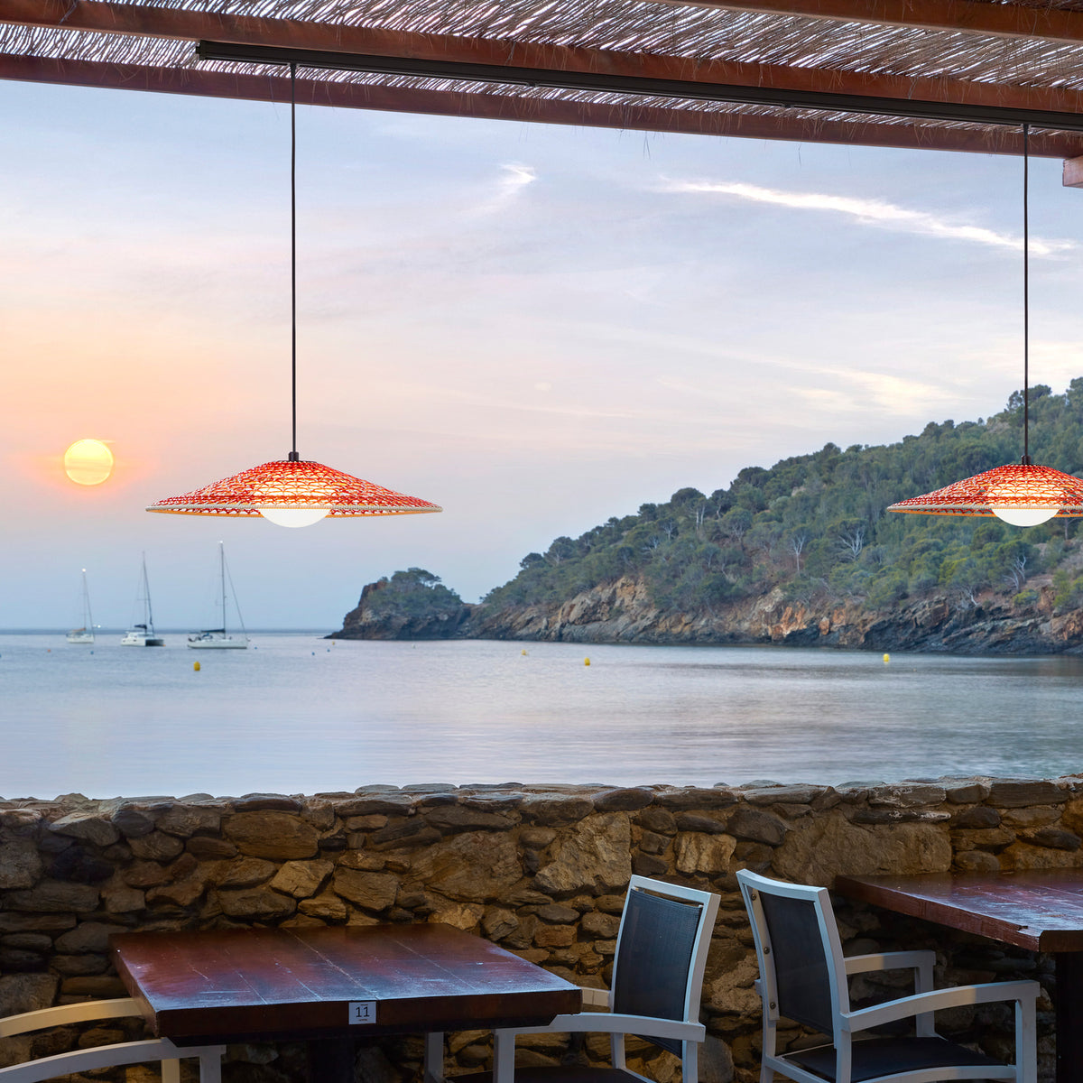 Nans S/55 Outrack woven LED outdoor track pendant lights with wide red woven shades and white diffusers, shown suspended above a coastal terrace dining area at sunset.