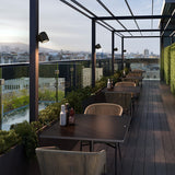 Nut A/01 Stainless Steel LED Outdoor Wall Light with rounded bell-shaped head on a slim rectangular plate, shown mounted along a rooftop terrace pergola at dusk.