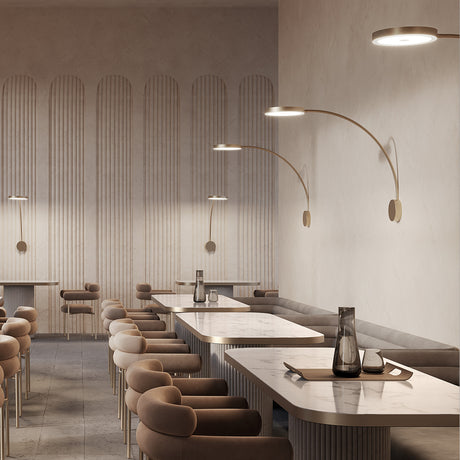 Dip Disc LED Wall Light in brushed brass with curved arm and round disc head, shown above marble dining tables in a modern restaurant setting.