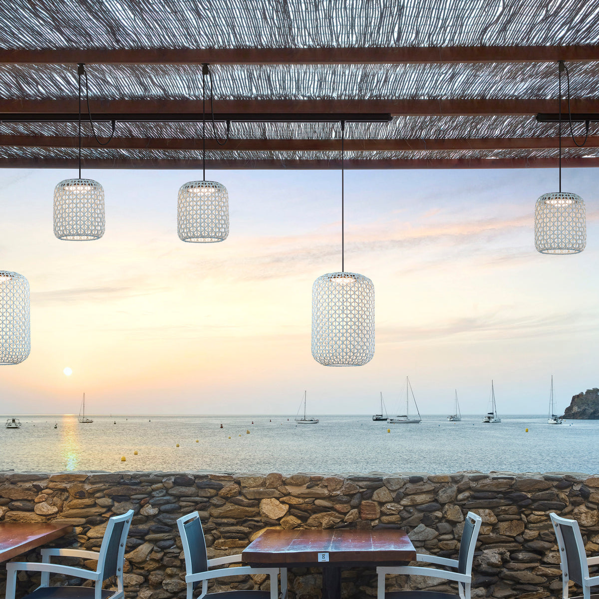Nans S/16 Outrack woven LED outdoor track pendants in beige shades on a track system, suspended over a coastal terrace dining area at sunset with sea views.