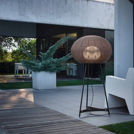 Garota P/02 outdoor floor lamp with a large woven brown globe shade on a slim three-legged graphite stand, shown illuminated on a patio beside a planter and outdoor seating.