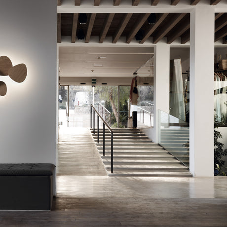 Tria 8 Light Wood LED Wall Light with overlapping pebble-shaped wood panels, shown illuminated beside a staircase in a modern interior setting.