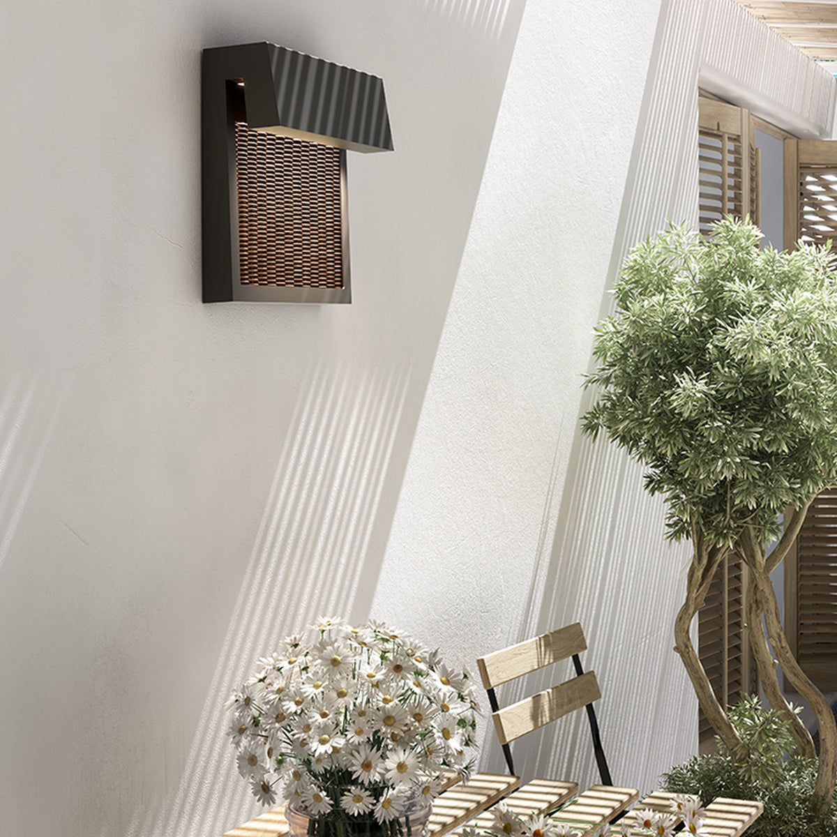 Sisal A/01 LED outdoor wall light with woven brown fibre front panel and angled hood, mounted beside a sunlit patio wall near plants and a wooden chair.