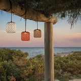 Nans S/16 woven outdoor pendant light with a brown cylindrical fabric shade, shown lit among white and red matching pendants hanging from a wooden beam at dusk by the sea.