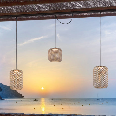 Nans S/31 Outrack LED outdoor track pendant lights with beige woven barrel-shaped shades, shown suspended from a pergola at sunset by the sea.