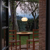 Garota S/01 Globe LED Outdoor Pendant Light in Ivory White, ribbed round shade glowing warmly above an outdoor dining table on a patio surrounded by trees and hedges.