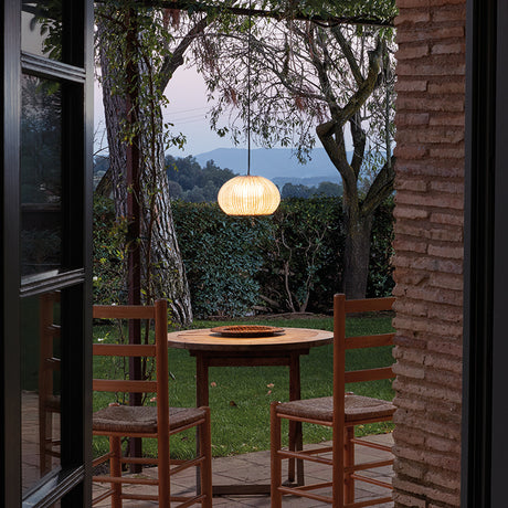 Garota S/01 Globe LED Outdoor Pendant Light in Ivory White, ribbed round shade glowing warmly above an outdoor dining table on a patio surrounded by trees and hedges.