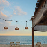 Garota S/01 Globe LED Outdoor Pendant Lights in brown woven shades, hung in a row from a beachside pergola overlooking the sea.