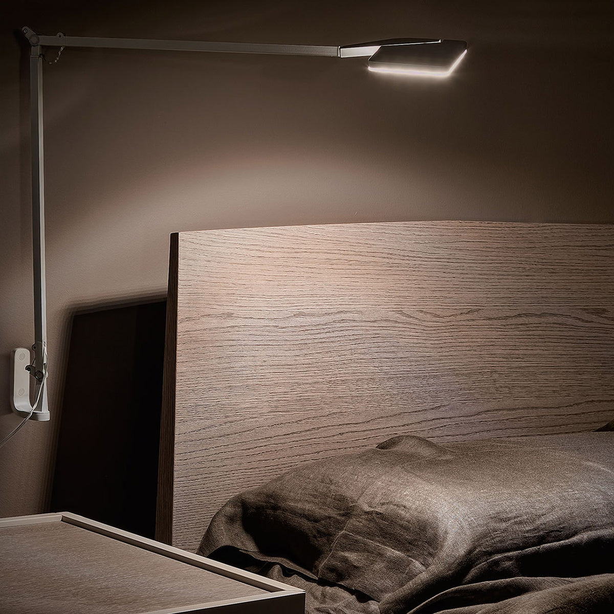 Jackie Aluminium LED Plug-in Wall Light with a long jointed arm and flat square head in white, mounted above a bed to cast warm ambient light over a wood headboard.