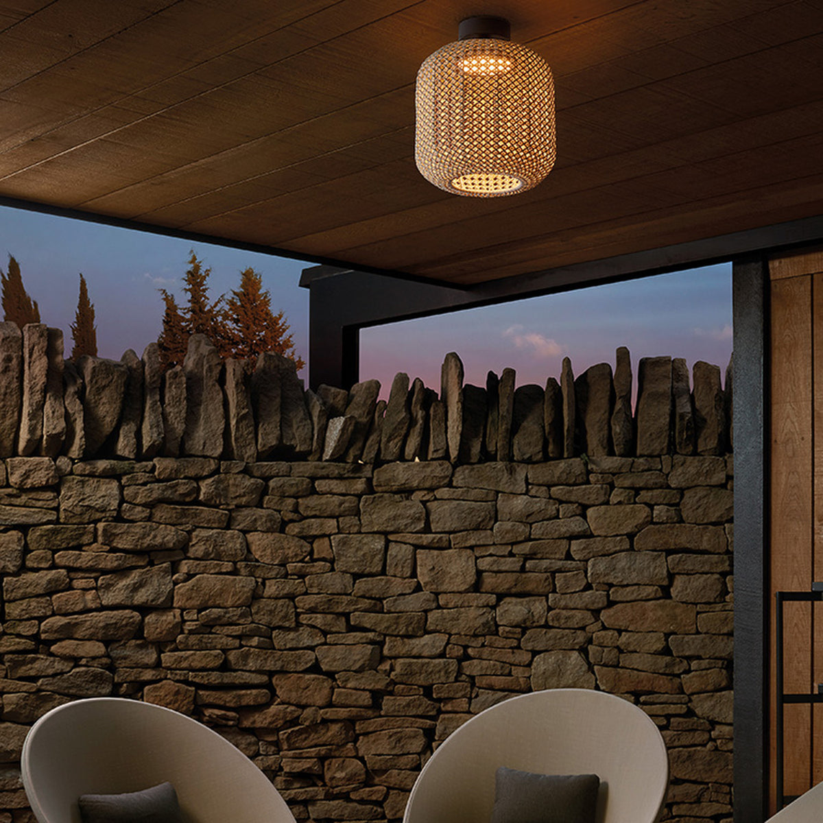 Nans PF/31 woven outdoor ceiling light with barrel-shaped beige fabric shade and dark mount, installed on a wooden patio ceiling above a stone wall.