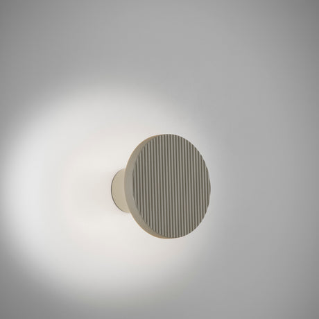 Arid A/01 round aluminium LED wall light with vertical fluted disc face in Textured Pebble Grey, mounted on a short cylindrical stem, shown glowing against a plain light grey wall.