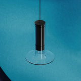 Auriel Glass Candle LED Pendant Light in matt black, a slim cylinder inside a clear glass tube with a flat glass disc base, suspended on a fine cable against a blue background.