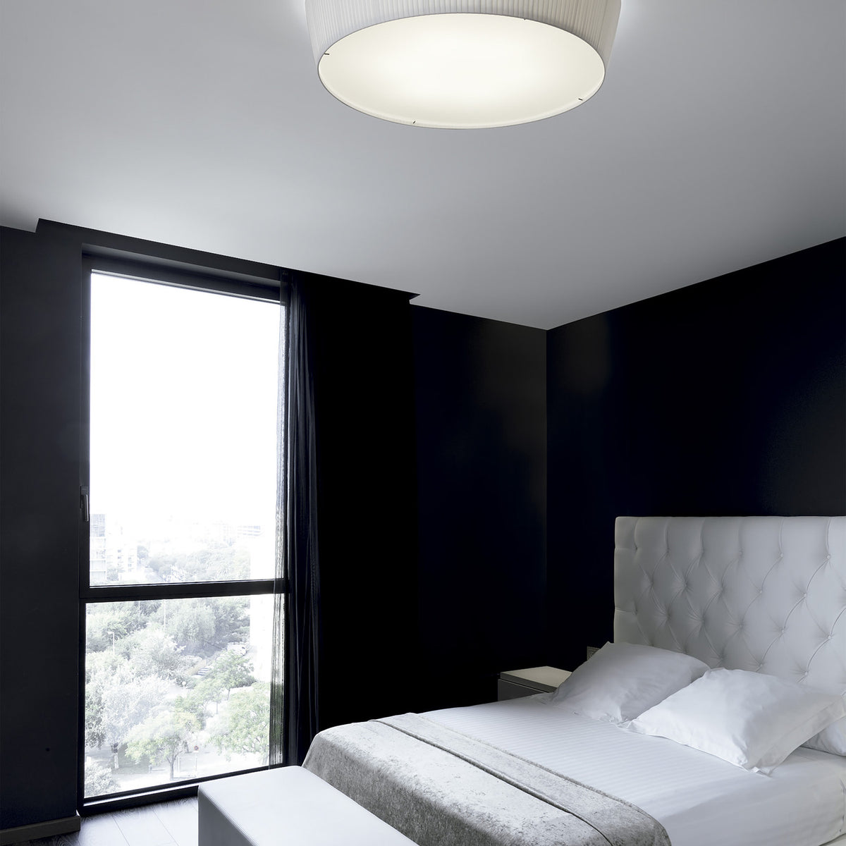 Plafonet 60 Ribbon LED ceiling light with a wide, shallow drum shade in pleated white translucent fabric, shown flush-mounted above a bed in a black-walled bedroom.