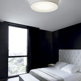 Plafonet 60 Ribbon LED ceiling light with a wide, shallow drum shade in pleated white translucent fabric, shown flush-mounted above a bed in a black-walled bedroom.